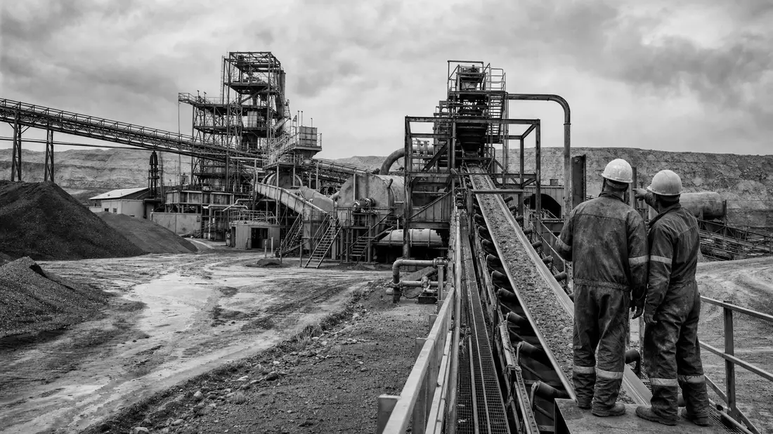 Schwarzweißes Reportagefoto von zwei Arbeitern an einer industriellen Förderbandanlage