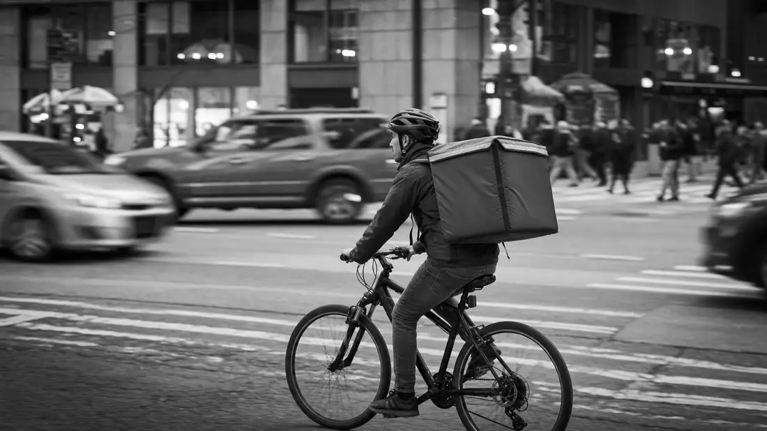 Dokumentarische Schwarzweißaufnahme eines Lieferfahrers mit Fahrrad im urbanen Straßenverkehr