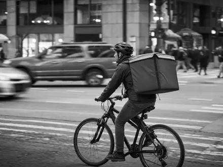 Schwarzweiß-Reportage dokumentiert Uber Technologies (US90353T1007): Essenslieferant auf Fahrrad mit unbedrucktem Lieferrucksack im Stadtverkehr - Foto: THN