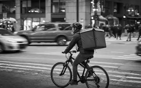 Schwarzweiß-Reportage dokumentiert Uber Technologies (US90353T1007): Essenslieferant auf Fahrrad mit unbedrucktem Lieferrucksack im Stadtverkehr - Foto: THN
