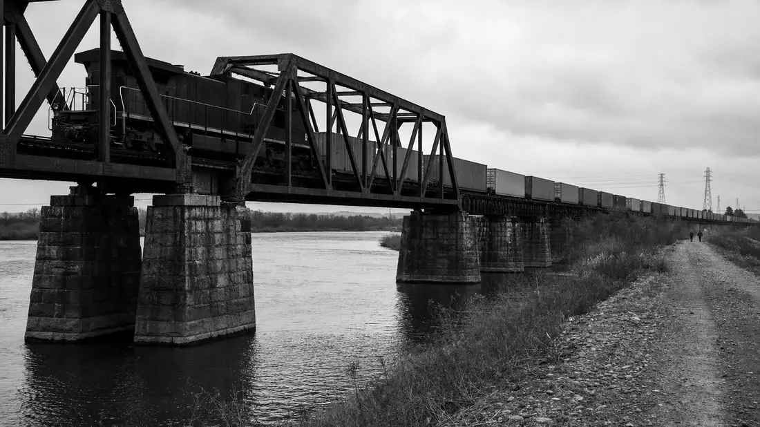 Schwarzweißes Dokumentarfoto eines Güterzugs auf einer Stahlbrücke über einen Fluss