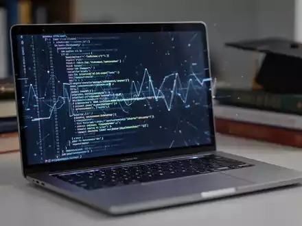 Nahaufnahme eines modernen silbernen Laptops mit leuchtendem Bildschirm, umgeben von verschwommenen Bildungsgegenständen, die Technologie und Bildung symbolisieren. - Foto: über boerse-global.de
