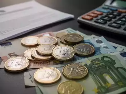 Euro-Münzen und -Banknoten liegen auf einer dunklen Oberfläche, im Hintergrund sind ein Taschenrechner und ein Dokument verschwommen zu sehen. - Foto: über boerse-global.de