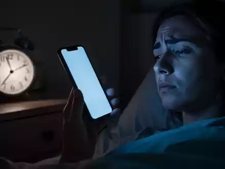Eine Person hält in einem dunklen Raum ein Smartphone, dessen bläuliches Licht das Gesicht beleuchtet. Eine Uhr zeigt eine späte Stunde an. - Foto: über boerse-global.de