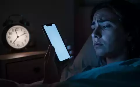 Eine Person hält in einem dunklen Raum ein Smartphone, dessen bläuliches Licht das Gesicht beleuchtet. Eine Uhr zeigt eine späte Stunde an. - Foto: über boerse-global.de