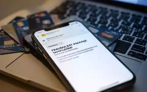 Ein Smartphone-Bildschirm zeigt eine betrügerische Paketbenachrichtigung, im Hintergrund verschwommen Kreditkarten und eine Laptoptastatur. - Foto: über boerse-global.de