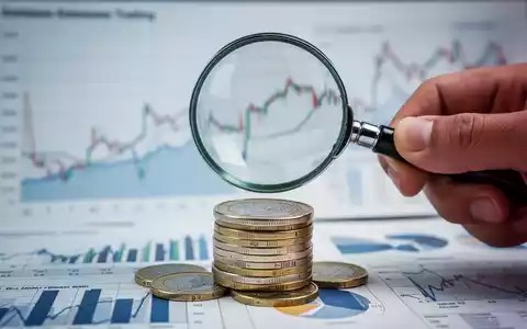 Eine Hand hĂ€lt eine Lupe ĂŒber einen Stapel Euro-MĂŒnzen, im Hintergrund verschwommene Diagramme, die finanzielle Spekulation darstellen. - Foto: ĂŒber boerse-global.de