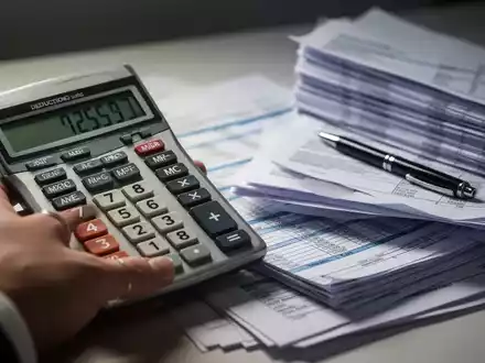Eine Nahaufnahme einer Hand, die einen Taschenrechner mit Zahlen auf dem Display hĂ€lt, neben einem Stapel Finanzdokumente und einem Stift. - Foto: ĂŒber boerse-global.de
