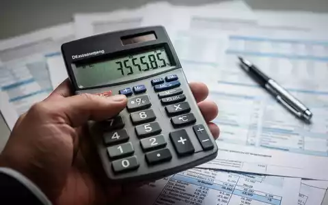 Eine Nahaufnahme einer Hand, die einen Taschenrechner hält, mit unscharfen Finanzdokumenten und einem Stift im Hintergrund. - Foto: über boerse-global.de
