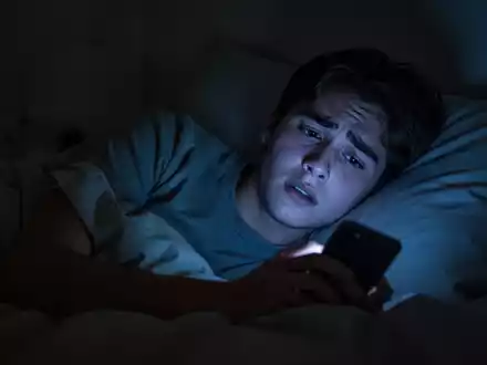 Eine junge Person liegt nachts wach im Bett, das Gesicht von einem Smartphone beleuchtet, das Sorgen und Schlaflosigkeit andeutet. - Foto: über boerse-global.de
