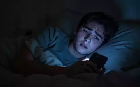 Eine junge Person liegt nachts wach im Bett, das Gesicht von einem Smartphone beleuchtet, das Sorgen und Schlaflosigkeit andeutet. - Foto: über boerse-global.de