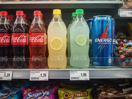 Nahaufnahme von zuckerhaltigen Getränken und Snacks im Supermarktregal mit Preisschildern, die eine Zuckersteuer andeuten. - Foto: über boerse-global.de