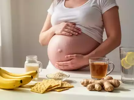 Eine schwangere Frau hält ihren Bauch, umgeben von Lebensmitteln wie Bananen, Reiskeksen, Ingwertee und Zitronenwasser. - Foto: über boerse-global.de