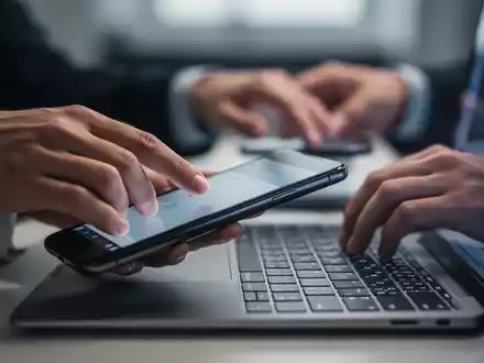 Eine Hand greift nach einem Smartphone, während eine andere versucht, sich auf eine Laptoptastatur zu konzentrieren, im Hintergrund verschwommen. - Foto: über boerse-global.de