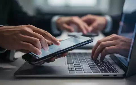 Eine Hand greift nach einem Smartphone, während eine andere versucht, sich auf eine Laptoptastatur zu konzentrieren, im Hintergrund verschwommen. - Foto: über boerse-global.de