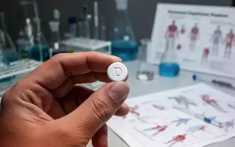 Eine Hand hält eine kleine weiße Vitamin-D-Pille vor einem unscharfen Hintergrund aus Laborgeräten und medizinischen Diagrammen. - Foto: über boerse-global.de