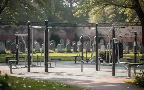 Ein moderner Outdoor-Fitnesspark mit Trainingsgeräten, eingebettet in eine grüne Parklandschaft, mit angedeuteten Grabsteinen im Hintergrund. - Foto: über boerse-global.de