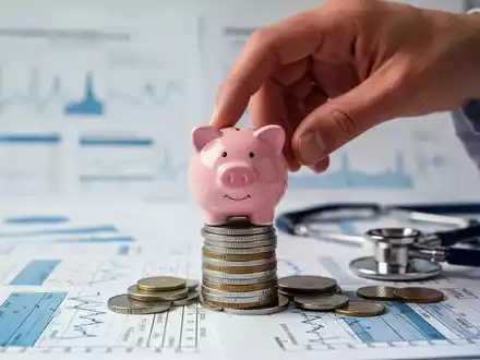 Eine Hand platziert ein Sparschwein auf Münzen vor verschwommenen medizinischen Diagrammen und einem Stethoskop. - Foto: über boerse-global.de