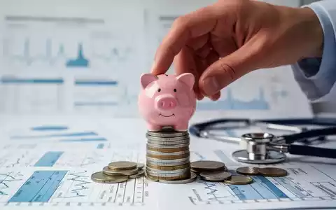 Eine Hand platziert ein Sparschwein auf Münzen vor verschwommenen medizinischen Diagrammen und einem Stethoskop. - Foto: über boerse-global.de