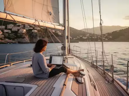 Eine Person arbeitet auf einem Laptop an Deck einer Segelyacht, mit dem Mittelmeer und einer Küste im Hintergrund. - Foto: über boerse-global.de