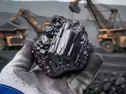 Nahaufnahme einer Graphitprobe, die in einer behandschuhten Hand vor einem unscharfen Bergbauhintergrund gehalten wird. - Foto: ĂŒber boerse-global.de