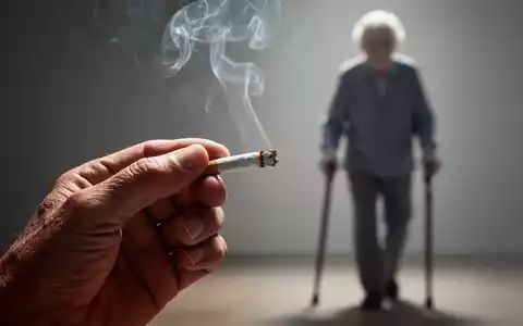Nahaufnahme einer Hand, die eine Zigarette hĂ€lt, mit Rauch, der aufsteigt. Im Hintergrund unscharf eine Ă€ltere Person mit Stock. - Foto: ĂŒber boerse-global.de