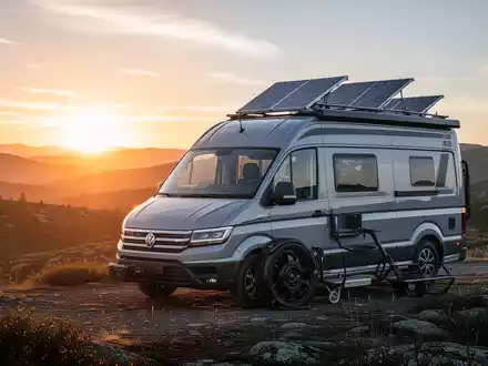 Ein moderner Camper Van, geparkt in einer malerischen Naturlandschaft bei Sonnenaufgang, mit sichtbaren Solarpaneelen auf dem Dach. - Foto: über boerse-global.de