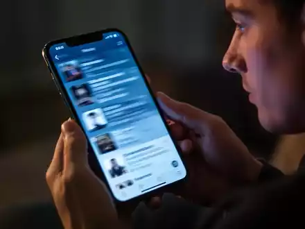 Eine Nahaufnahme eines Smartphones, das in Händen gehalten wird, der Bildschirm zeigt einen schnell scrollenden Feed in einem abgedunkelten Raum. - Foto: über boerse-global.de