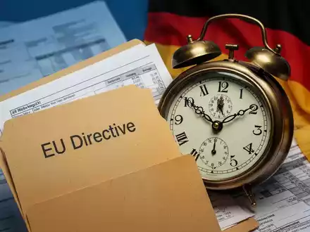 Eine leicht geöffnete Akte mit der Aufschrift 'EU-Richtlinie' und eine Uhr, die eine verstrichene Frist anzeigt, mit deutscher Flagge im Hintergrund. - Foto: über boerse-global.de
