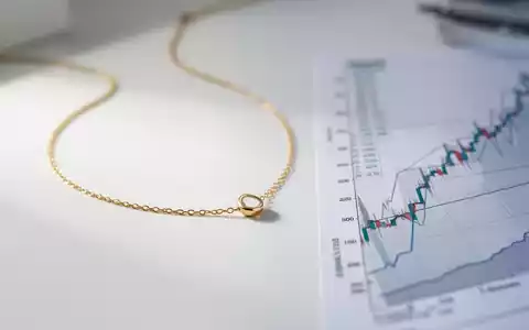 Eine filigrane goldene Halskette liegt auf einer sauberen weißen Oberfläche, daneben eine unscharfe Finanzgrafik mit Aufwärtstrend. - Foto: über boerse-global.de