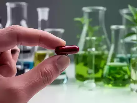 Eine Hand hält eine rote Berberin-Kapsel über einem unscharfen Hintergrund mit Laborgeräten und Pflanzenextrakten. - Foto: über boerse-global.de