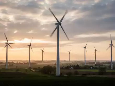 Ein Windpark in einer ländlichen deutschen Landschaft bei Sonnenaufgang oder Sonnenuntergang, mit modernen Windturbinen vor einem dramatischen Himmel. - Foto: über boerse-global.de