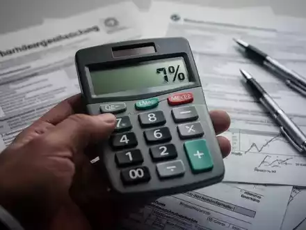Eine Hand hält einen Taschenrechner, der einen negativen Prozentsatz anzeigt, mit verschwommenen Steuerdokumenten im Hintergrund. - Foto: über boerse-global.de