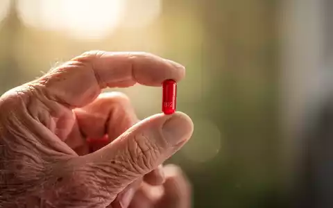 Eine leicht faltige Hand hält eine rote Kapsel, die Vitamin B12 symbolisiert, vor einem weichen, warmen Hintergrund. - Foto: über boerse-global.de