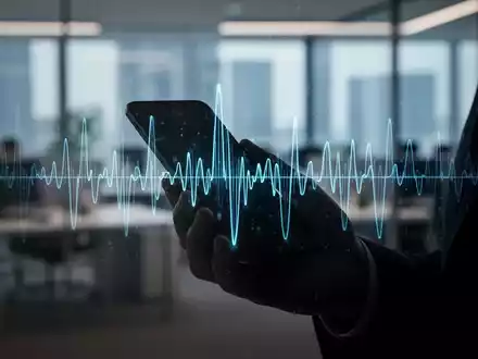 Eine Hand hĂ€lt ein Smartphone, aus dem eine KI-generierte Sprachwellenform abstrahlt, vor einem unscharfen BĂŒrohintergrund. - Foto: ĂŒber boerse-global.de