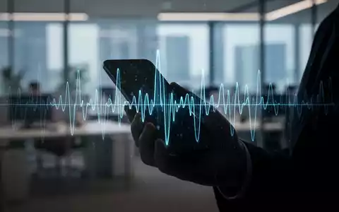 Eine Hand hält ein Smartphone, aus dem eine KI-generierte Sprachwellenform abstrahlt, vor einem unscharfen Bürohintergrund. - Foto: über boerse-global.de