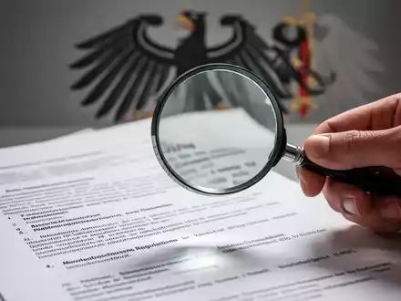 Eine Hand hält eine Lupe über ein Dokument, das Tarifverträge symbolisiert, im Hintergrund verschwommen ein Bundesadler. - Foto: über boerse-global.de