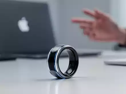 Futuristischer smarter Ring mit dezent leuchtenden Details, für Produktivität, liegt auf sauberer Fläche neben Apple-Laptop. - Foto: über boerse-global.de