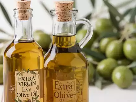 Nahaufnahme einer Glasflasche Olivenöl mit unscharfem Hintergrund aus frischen grünen Oliven und Blättern. - Foto: über boerse-global.de