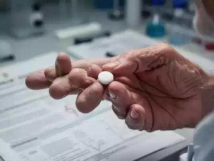 Eine Nahaufnahme der Hand einer älteren Person, die eine weiße Pille hält, mit einem unscharfen medizinischen Hintergrund. - Foto: über boerse-global.de
