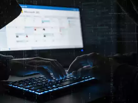 Schattenhafte Hände tippen auf Tastatur, verschwommener Microsoft 365-Kalender und digitale Code-Overlays, die Cyberbedrohungen darstellen. - Foto: über boerse-global.de