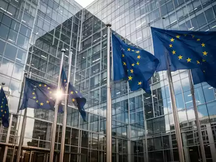 Europäische Flaggen vor einem modernen Bürogebäude als Symbol für regulatorische Eingriffe der EU-Kommission. - Foto: über boerse-global.de