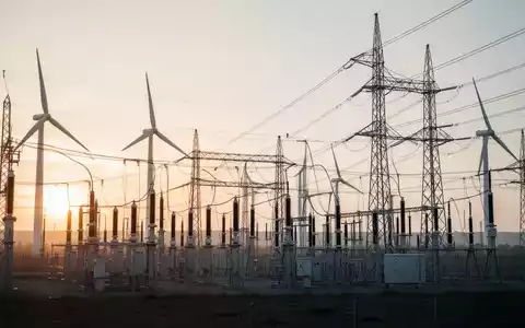 Moderne Hochspannungstrassen und Windkraftanlagen in einer technologisch geprägten Industrielandschaft bei Sonnenaufgang. - Foto: über boerse-global.de