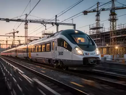 Ein moderner Regionalzug fährt in der Abenddämmerung an einer Baustelle mit Baugerüsten und Kränen entlang. - Foto: über boerse-global.de