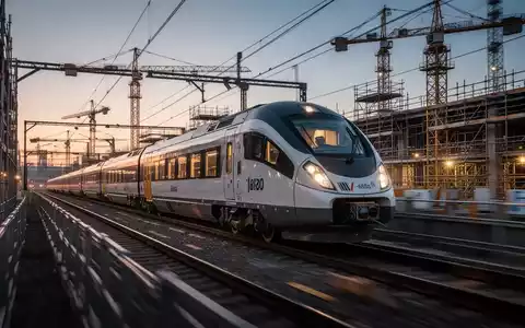 Ein moderner Regionalzug fährt in der Abenddämmerung an einer Baustelle mit Baugerüsten und Kränen entlang. - Foto: über boerse-global.de
