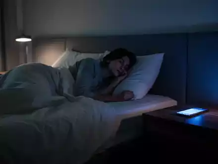 Eine Person liegt nachts in einem abgedunkelten Schlafzimmer, während das Licht eines Smartphones auf den Nachttisch fällt. - Foto: über boerse-global.de