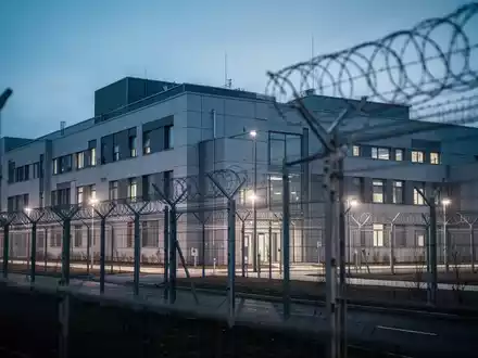 Modernes Gebäude einer forensischen Psychiatrie bei Dämmerung mit Sicherheitszaun und dezenter Beleuchtung. - Foto: über boerse-global.de