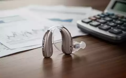 Ein modernes Hörgerät liegt neben einem Taschenrechner und Finanzunterlagen auf einem Holztisch. - Foto: über boerse-global.de