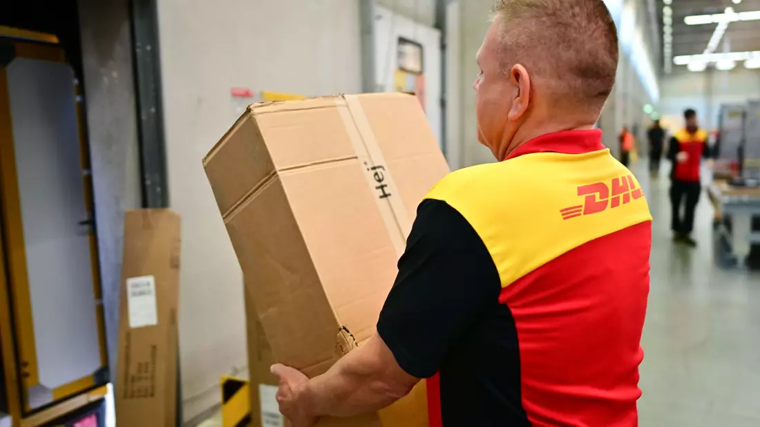 Ein Paketbote schleppt in einem DHL-Standort ein schweres Paket. (Archivbild) - Bild: Sebastian Christoph Gollnow/dpa