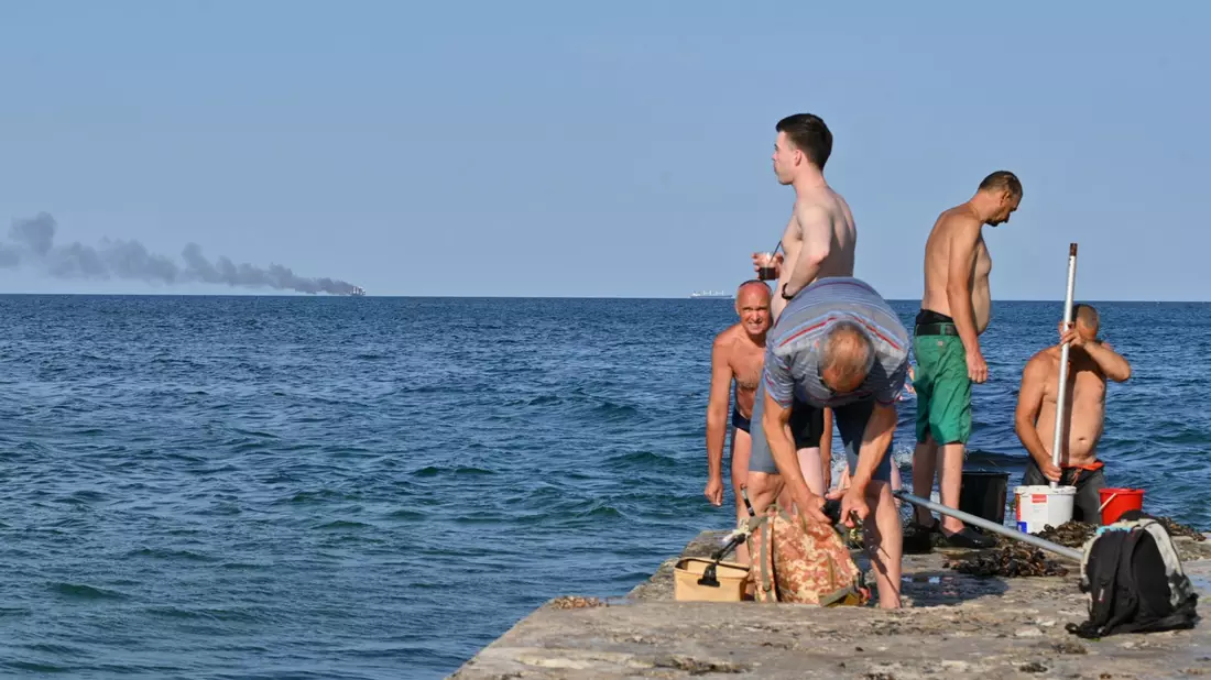 Menschen in Odessa sonnen sich, während am Horizont im Schwarzen Meer der brennende Getreidetransporter «Golden Leo» zu sehen ist. (Archivbild) - Bild: Michael Shtekel/AP/dpa